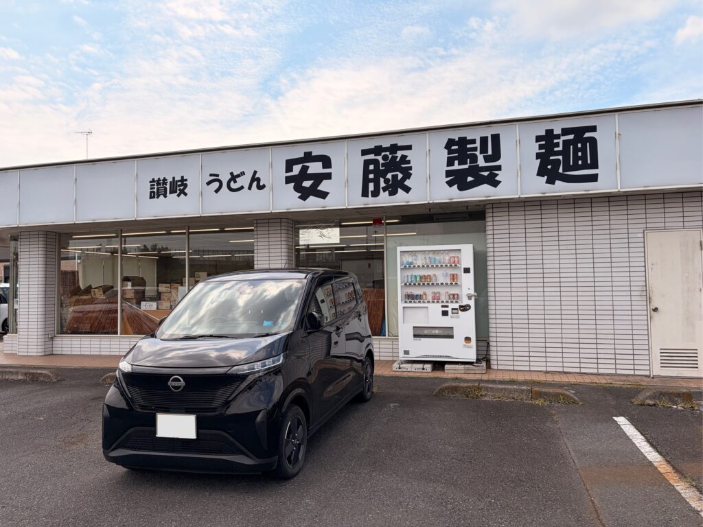 「讃岐うどん 安藤製麺」の店舗外観。看板の下に黒い日産サクラと自動販売機