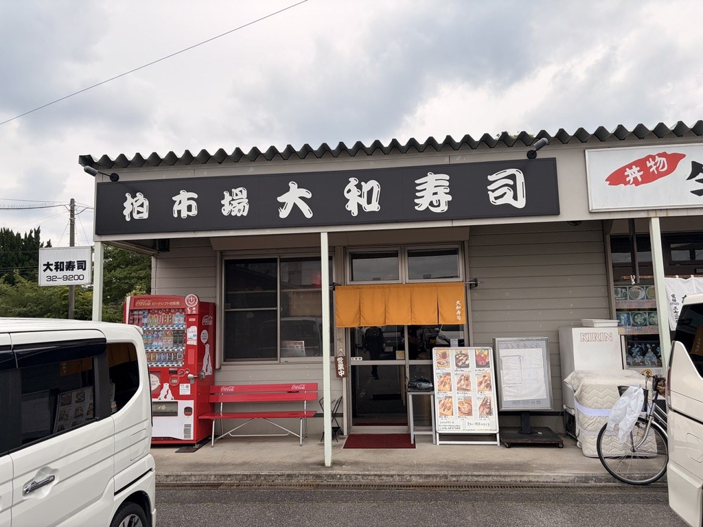 柏市場 大和寿司の外観。黄色い暖簾と看板、店頭の赤いベンチ